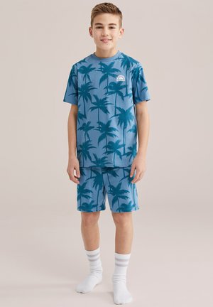 Fant, stoječ, oblečen v modro srajco z vzorcem palm in ujemajoče kratke hlače, bele nogavice, na preprosti svetli podlagi.