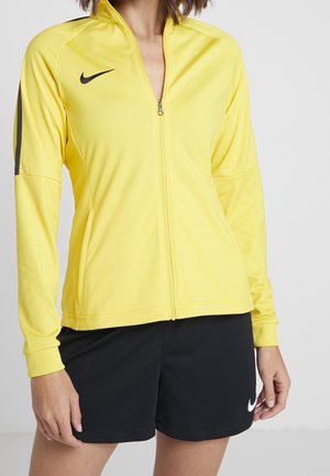 Femme portant une veste sportive jaune vif à fermeture éclair et un short noir Nike, debout devant un fond clair uni.