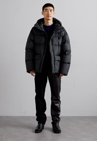 Chaqueta acolchada negra con capucha, con un diseño acolchado y cierre de cremallera. Se lleva sobre una camiseta negra y combinada con pantalones oscuros y botas.