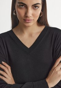 Pull noir en V, texture douce, encolure et poignets côtelés, porté les bras croisés, mettant en valeur un gros plan du vêtement.