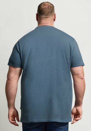 Uomo con capelli corti che indossa una maglietta blu tinta unita a maniche corte, visto di spalle su uno sfondo chiaro uniforme.