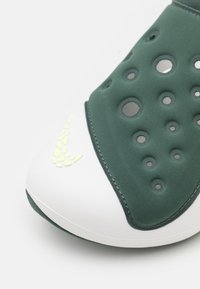 Grön och vit sko med en slät, strukturerad ovandel med ventilationshål, samt en gul accent nära tån. Kurvad, modern design.