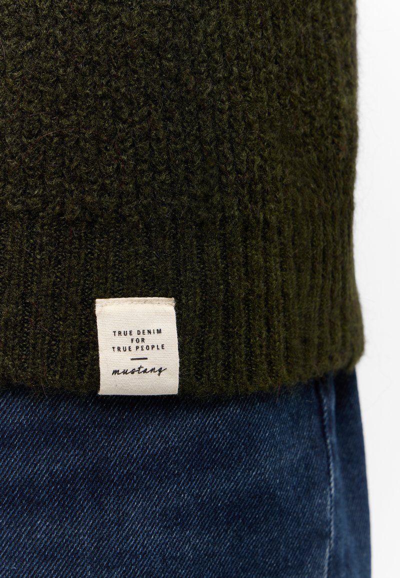 Doce de un suéter de punto verde oscuro con una etiqueta de tela blanca que dice "Denim verdadero para gente verdadera" sobre unos jeans de mezclilla azul.