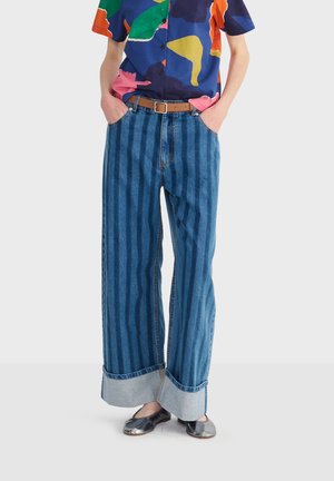 Osoba oblečená v širokých modrých rifliach s vertikálnymi pruhmi, farebnej abstraktnej košeli na gombíky, hnedom opasku a metalických balerínach.