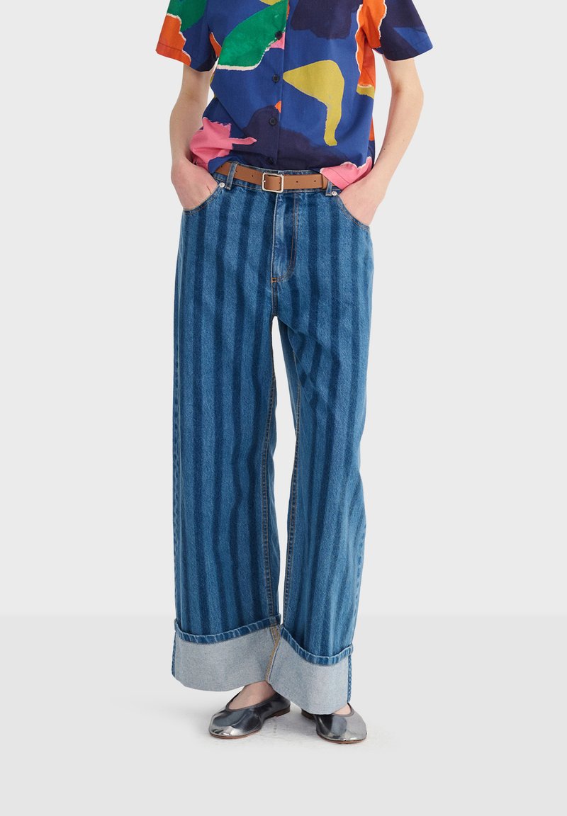 Person trägt weit geschnittene blaue Jeans mit vertikalen Streifen, ein buntes abstraktes Hemd zum Knöpfen, einen braunen Gürtel und metallische Ballerinas.