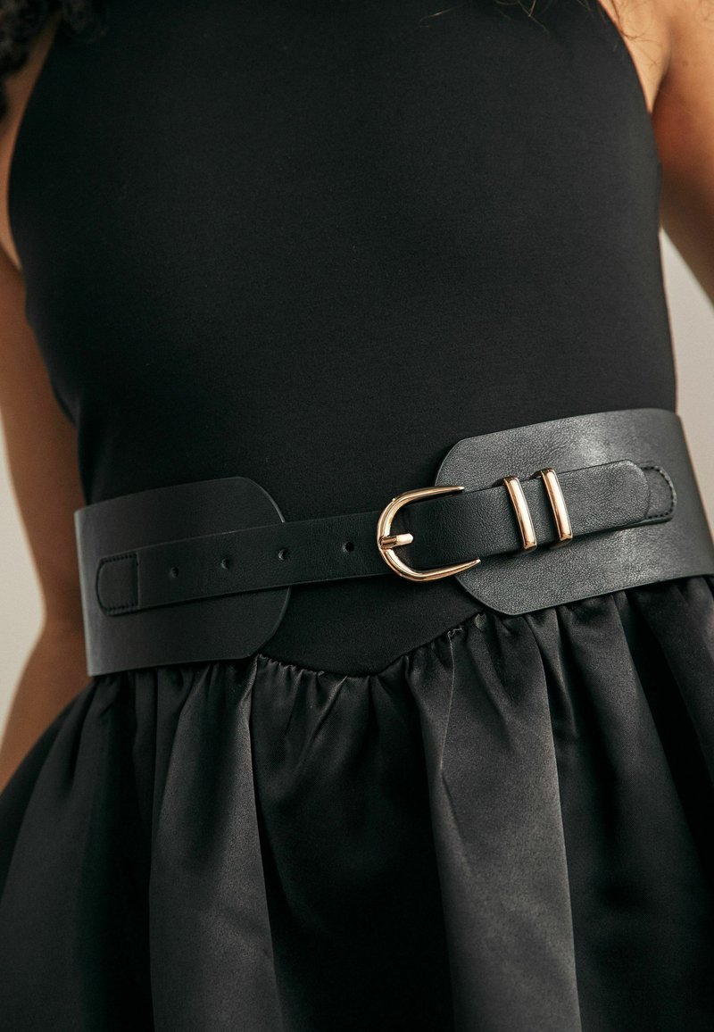 Black leather belt with a wide design, featuring a gold metal buckle and two parallel slots. Paired with a black, gathered fabric dress.