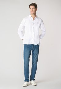 Camicia bianca di lino con colletto a bottoni, maniche lunghe arrotolate ai polsini, abbinata a jeans blu e sneakers bianche. Presenta un piccolo logo.