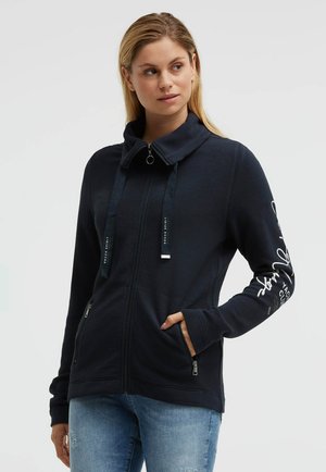 Woman wearing a navy zip-up jacket with text on the right sleeve, hands in pockets, looking slightly to the side, against a plain background.