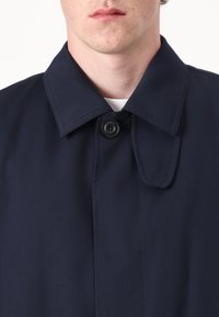Mørkeblå frakk med strukturert krage, som har en enkelt svart knapplås og glatt tekstur. Minimalistisk design uten synlige mønstre.