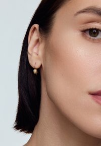 Złota kolczyk w kształcie obręczy z teksturowanym, okrągłym charms. Zbliżenie na kolczyk przyczepiony do ucha, z gładką skórą i widocznymi ciemnymi włosami.