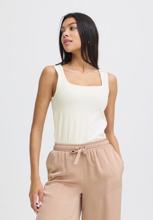 Jeune femme aux longs cheveux noirs portant un haut blanc sans manches et un pantalon beige à cordon, debout avec une main dans la poche.