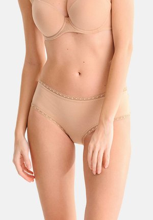 Femme portant un soutien-gorge beige et un slip taille haute assorti avec bordure en dentelle sur un fond uni clair.