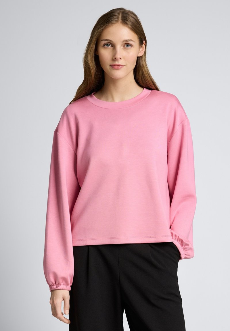 Frau mit langen braunen Haaren, die ein lockeres, rosa Langarmshirt und schwarze Hosen trägt, vor einem schlichten grauen Hintergrund steht.