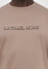 Lys beige T-skjorte laget av myk bomull, med en hevet Michael Kors-logo i en mørkere nyanse på brystet. Rund hals.