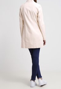 Abrigo largo beige claro con una textura suave, diseño ajustado y cuello alto; combinado con jeans ajustados azul oscuro y zapatillas blancas.