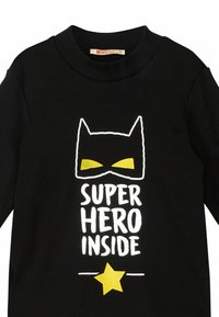 Camicia nera di cotone a maniche lunghe con il testo bianco "SUPER HERO INSIDE", stella gialla e grafica di una maschera da supereroe gialla e nera.