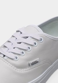 Λευκό sneaker Vans με λευκά κορδόνια, μεταλλικά ροδέλες και υφασμάτινη ετικέτα που δείχνει το λογότυπο της μάρκας Vans σε δερμάτινο επάνω μέρος.