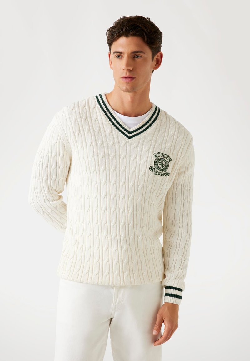 Fehér cable-knit pulóver V-nyakkivágással, sötétzöld és fehér csíkos díszítéssel a galléron és az ujjakon, valamint zöld hímzett logóval.