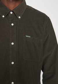Dark green corduroy shirt featuring a button-down collar, front placket with buttons, and a left chest pocket with a branded label.