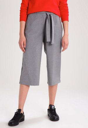 Femme portant un pantalon gris large et court avec une ceinture nouée, un haut rouge à manches longues et des baskets noires, debout sur un fond blanc.