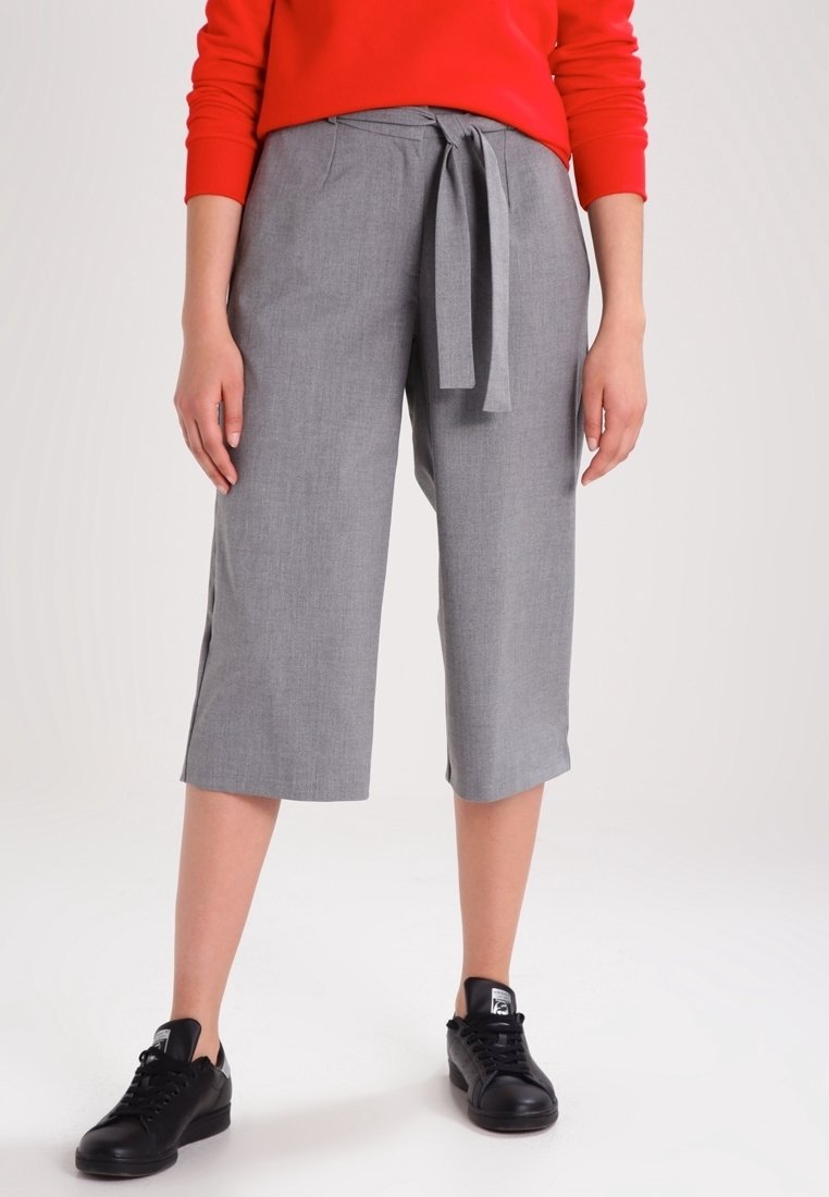 Femme portant un pantalon gris large et court avec une ceinture nouée, un haut rouge à manches longues et des baskets noires, debout sur un fond blanc.