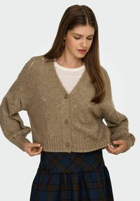 Beige strikket cardigan med V-hals, store knapper og udskårne mønstre. Bæres over en hvid top og lagdelt med et ternet nederdel.