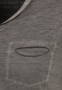 Camiseta de algodón gris con cuello redondo y un pequeño bolsillo en el pecho con una abertura redondeada, exhibida sobre una superficie lisa y texturizada.