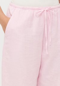 Pantaloni in lino rosa chiaro con cintura elastica e cordoncino. Presentano tasche laterali e una vestibilità rilassata. Tessuto liscio e texture.