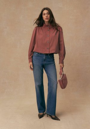 Frau mit langen braunen Haaren, die eine rostfarbene, geknöpfte Bluse, blaue Jeans, Leopardenmuster-High Heels trägt und eine passende rostfarbene Handtasche hält.