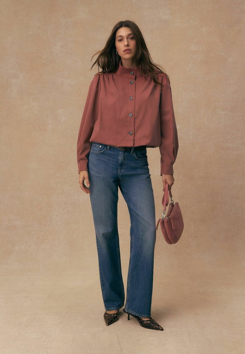 Femme aux longs cheveux bruns portant un chemisier rouille à boutons, un jean bleu, des talons imprimés léopard et tenant un sac à main assorti de couleur rouille.