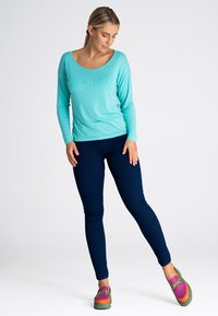 Długi rękaw, turkusowa bluzka z okrągłym dekoltem, zestawiona z granatowymi, obcisłymi dżinsami i wielokolorowymi klapkami z zieloną podeszwą.