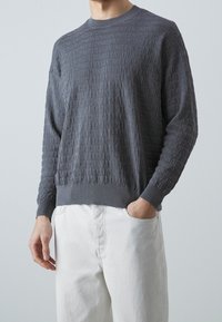 Szary sweter z wypukłym wzorem logo, z okrągłym dekoltem oraz ściągaczami przy mankietach i dole. Noszony z białymi spodniami, prezentując casualowe warstwowe ubranie.