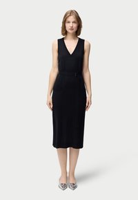 Svart ärmlös klänning med V-ringning, försedd med bälte, två framfickor och rak skärning. Modellen har på sig silverfärgade skor med spetsig tå.