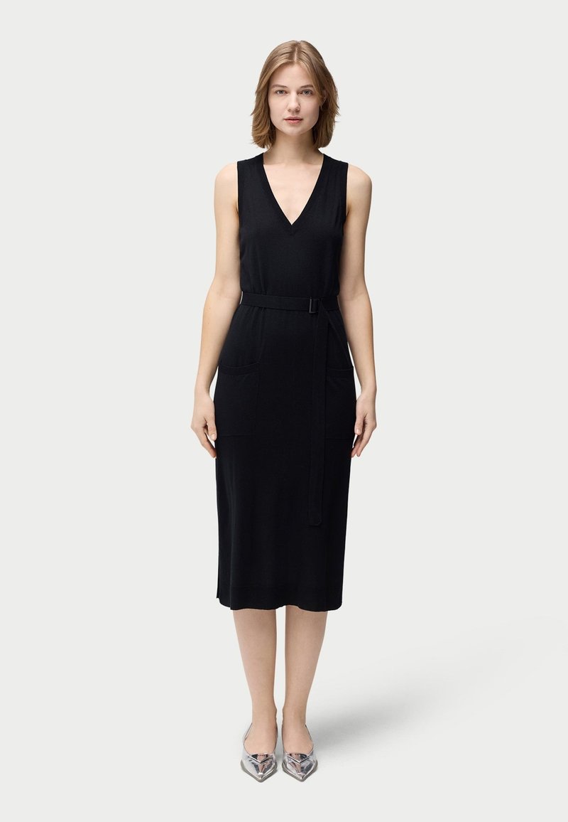Svart ärmlös klänning med V-ringning, försedd med bälte, två framfickor och rak skärning. Modellen har på sig silverfärgade skor med spetsig tå.