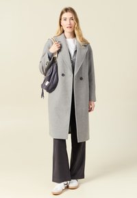 Grå ullrock med slagkrage och dubbelradiga knappar, bärs över en mörk outfit, kombinerad med en texturerad lila handväska.