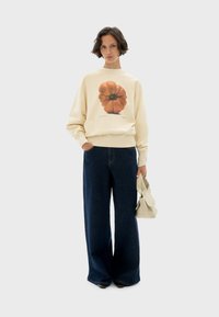 Cream sweatshirt featuring an orange pumpkin graphic, paired with wide-leg dark blue jeans and a light beige bag. Simple, casual design.