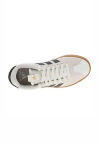 Zapatilla deportiva blanca con rayas negras, parte superior texturizada, suela de goma, cuello acolchado y cordones blancos. Presenta un logo dorado en la lengüeta.