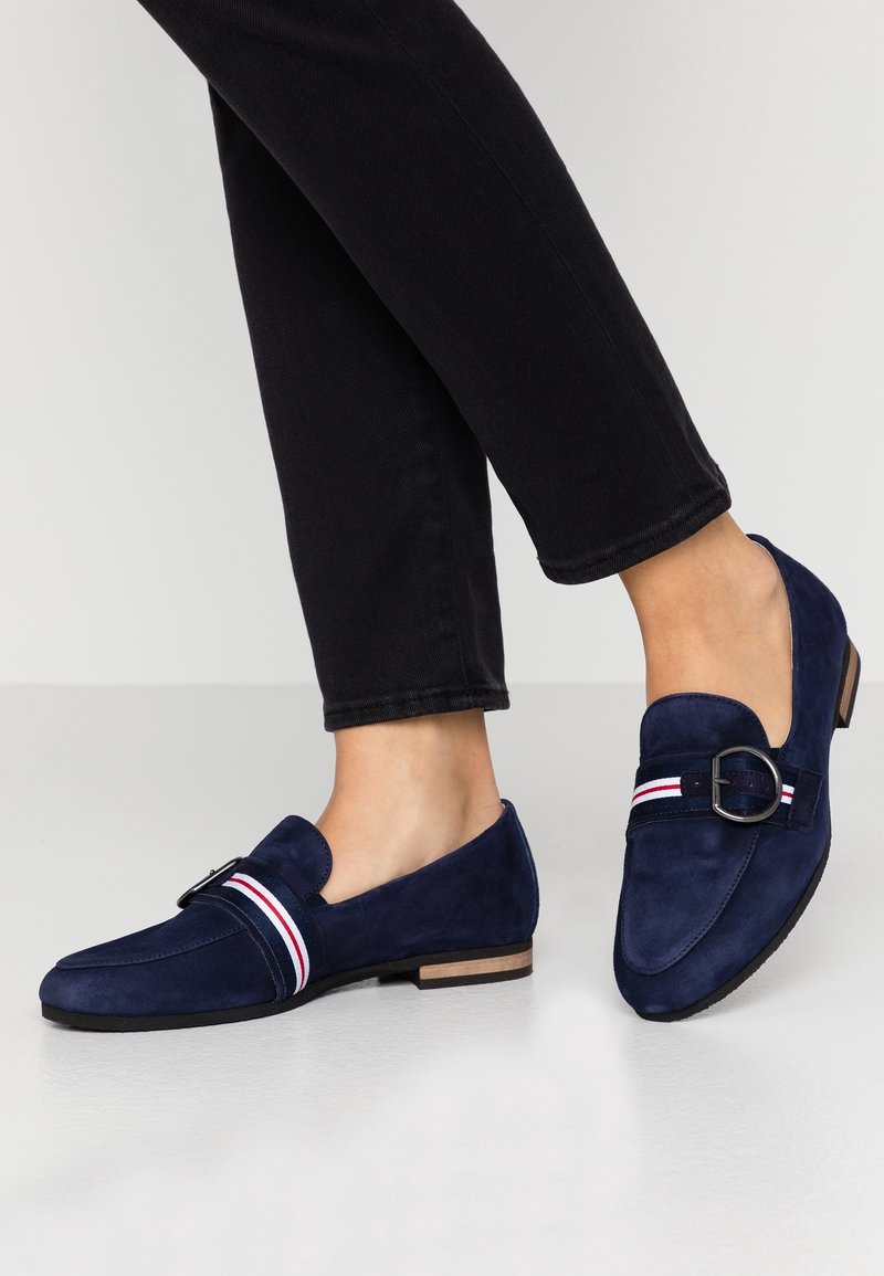 Pieds chaussés de mocassins en daim bleu marine avec une bride rayée rouge, blanche et bleue et une boucle en métal, associés à un pantalon noir.