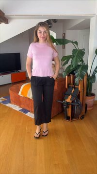 Νέα γυναίκα με ροζ τοπ και μαύρο παντελόνι στέκεται ξυπόλητη σε ένα σαλόνι με πορτοκαλί καναπέ, μεγάλο πράσινο φυτό και ξύλινο πάτωμα.