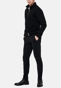 Mand iført sort quarter-zip sweater, sorte slim bukser med bælte og sorte læder ankelstøvler, stående med hænderne i lommerne.