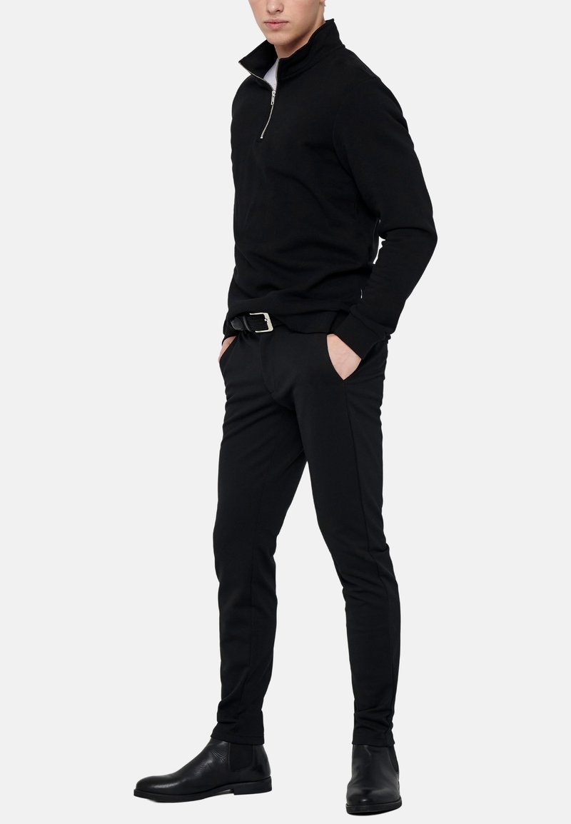 Mand iført sort quarter-zip sweater, sorte slim bukser med bælte og sorte læder ankelstøvler, stående med hænderne i lommerne.