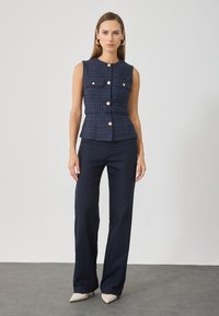Top fără mâneci din tweed albastru închis, cu talie ajustată, având patru buzunare frontale și butoane aurii. Asortat cu pantaloni navy cu talie înaltă.