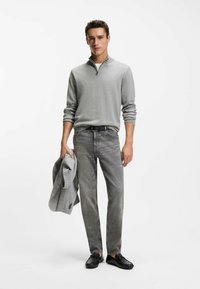 Szary sweter z suwakiem do ćwiartki, jasnoszare dżinsy i czarne buty wsuwane. Trzyma szarą marynarkę. Prosty wzór, miękka faktura, neutralne kolory.