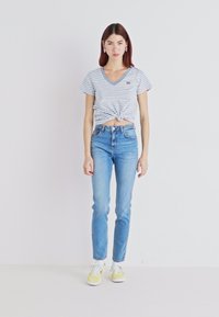 Top azul e branco de algodão, com riscas, de mangas curtas e decote em v com nó, combinado com jeans de cintura alta, em azul claro, e ténis amarelos.