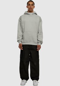 Sudadera gris con capucha y bolsillo canguro, combinada con pantalones cargo negros de corte holgado y zapatillas blancas, sobre un fondo neutro.