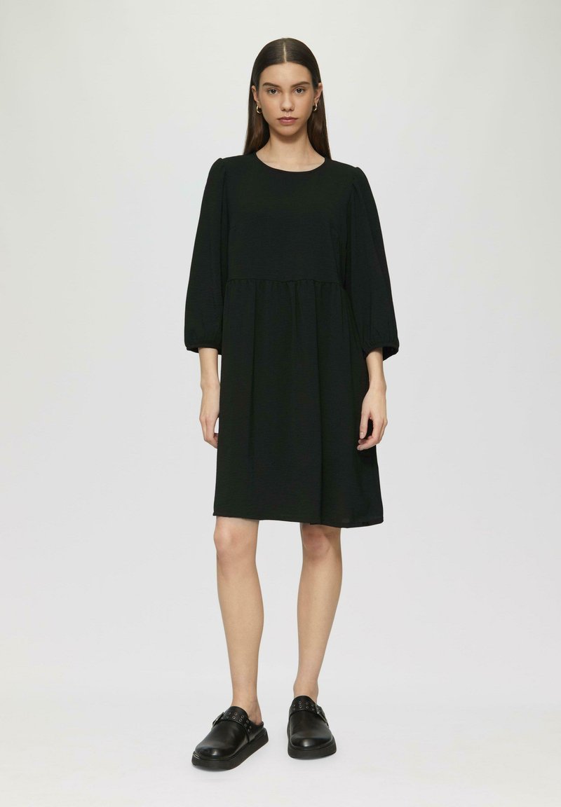Schwarzes Kleid mit Puffärmeln, rundem Ausschnitt, geraffter Taille und weicher Textur. Kombiniert mit schwarzen Slippern mit Riemenakzenten.