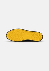 Semelle de chaussure noire avec un dessous en caoutchouc jaune texturé. Présente des motifs en zigzag et en losange pour une meilleure adhérence. Logo rond au centre.