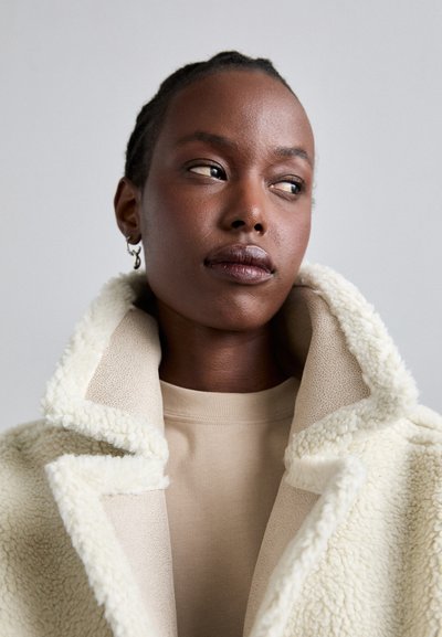 Witte, pluche, schapenvachtjas met een brede kraag, gelaagd over een beige top. Zachte textuur met een contrast tussen stoffen en kleuren.
