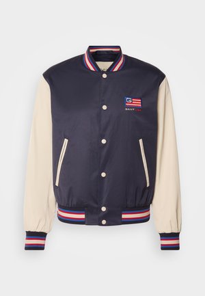 Marineblå varsityjakke med cremefarvede ærmer, stribede ribbede krave og manchetter, broderet logo og trykknapper. To sidelommer.