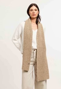 Scarf di maglia beige con una texture a coste, avvolta attorno al collo, abbinata a un maglione bianco e a pantaloni di colore chiaro.
